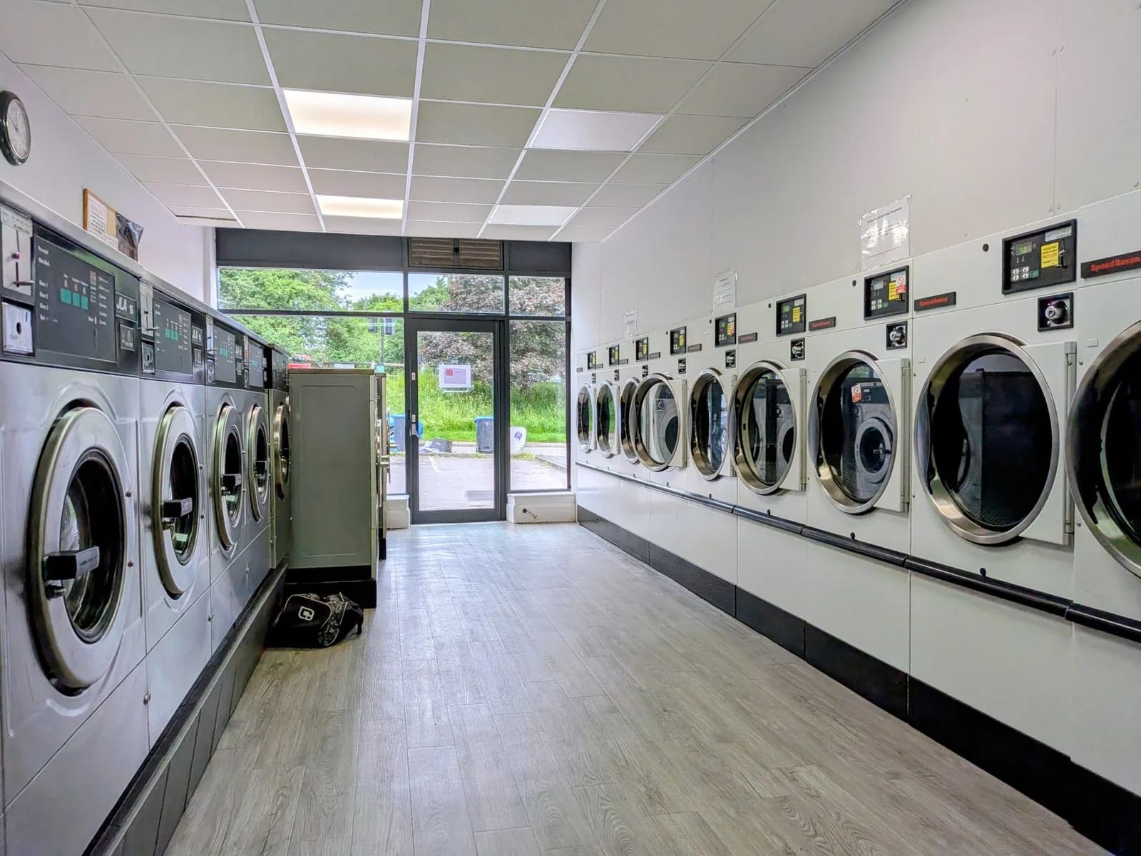 A narrow laundromat with front-loading washers along one wall and dryers along the other, and a glass door to the street