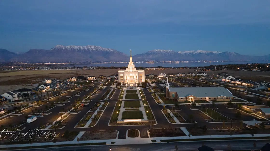 An aerial view of the Saratoga Springs Utah Temple, a light stone building with a tall central spire, on its landscaped grounds