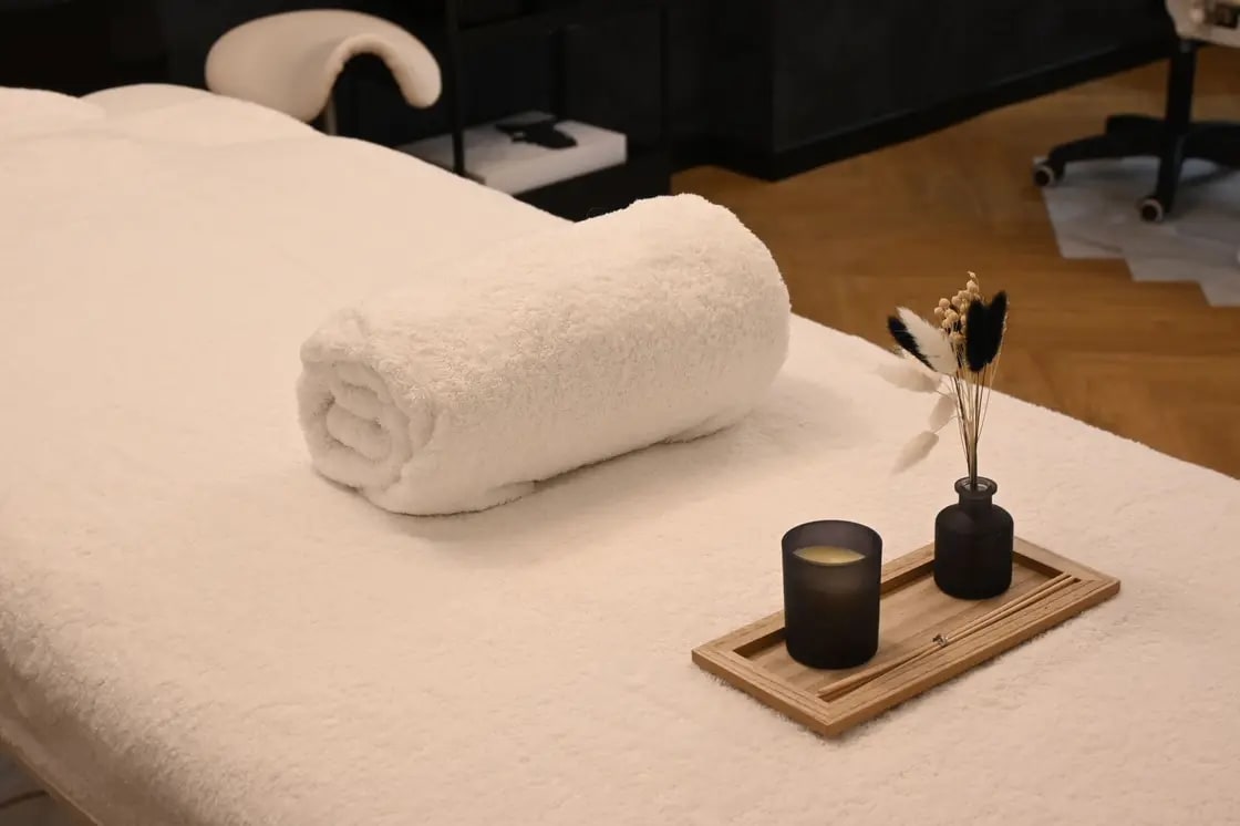 A rolled white towel and a small tray holding a candle and a dried-flower vase on a massage table