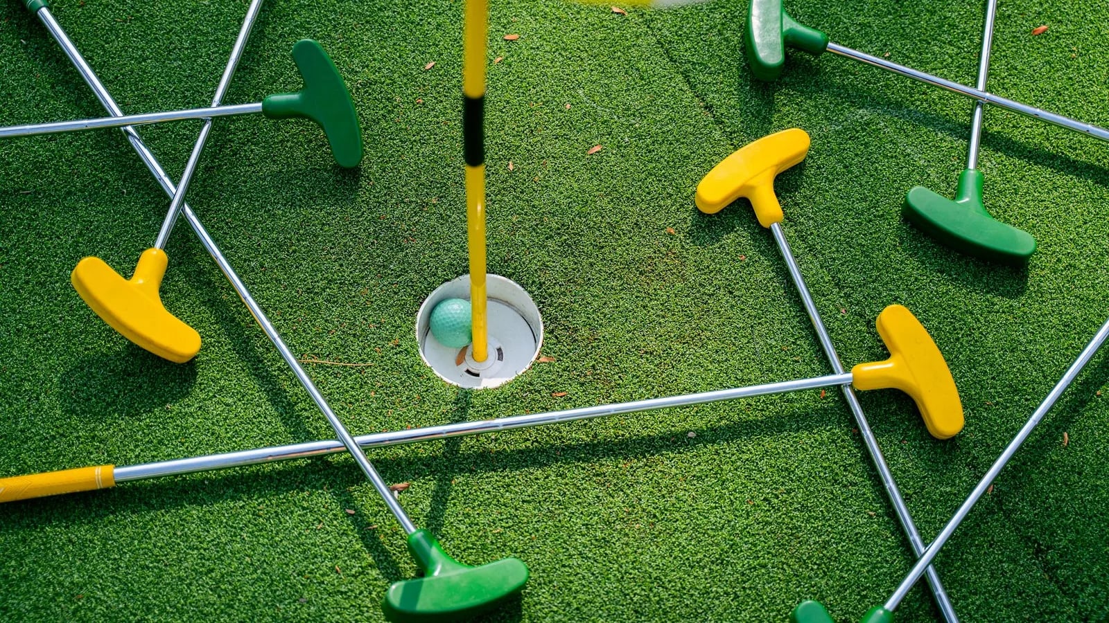 Brightly colored putters arranged around a cup on an artificial-turf mini golf green