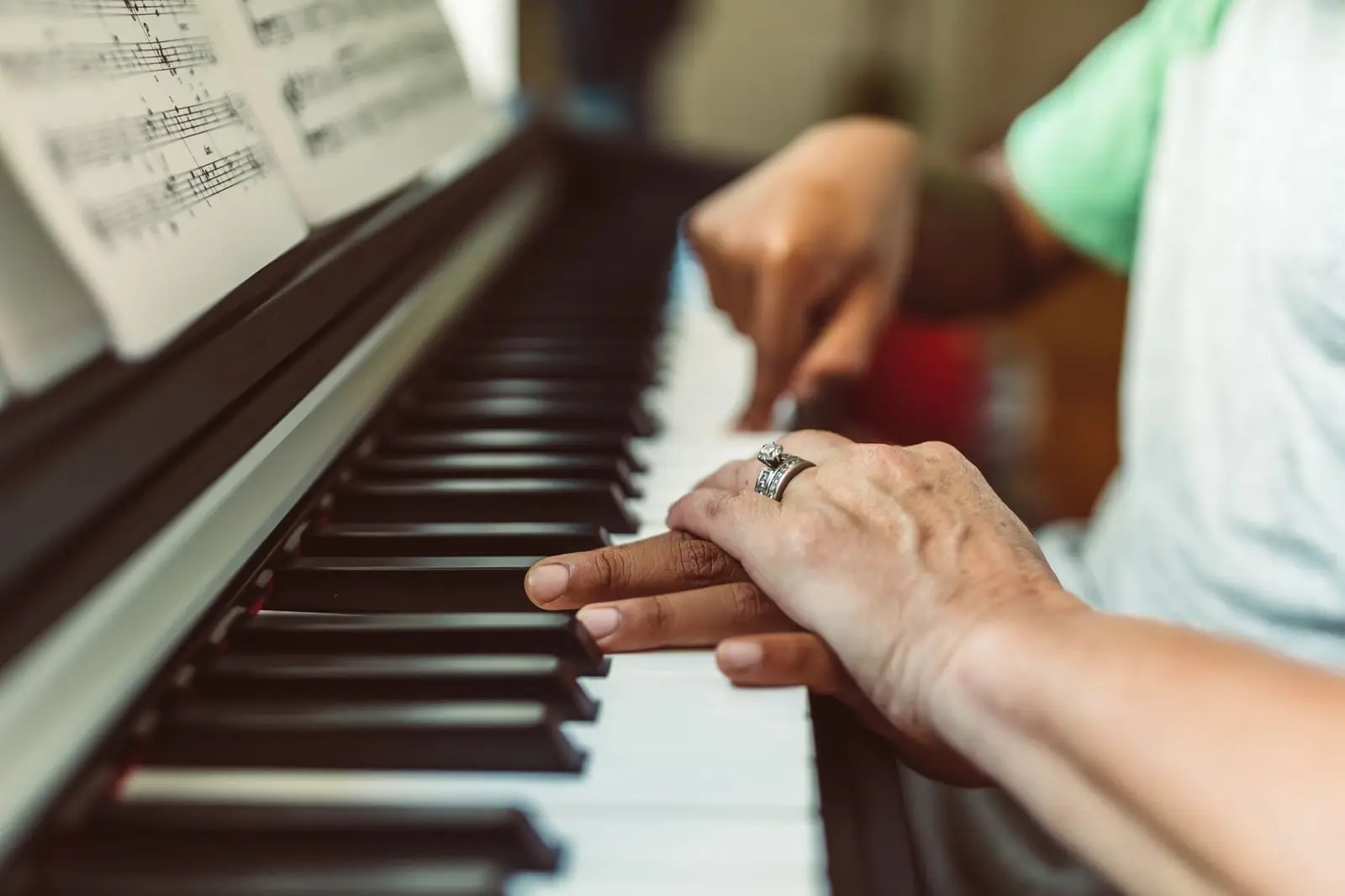 An older adult's hand resting over a younger hand at the keys of an upright piano, sheet music open on the stand