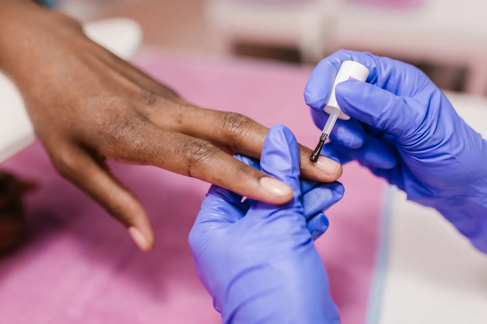 A technician in purple gloves applying polish to a client's fingernail at a pink salon table