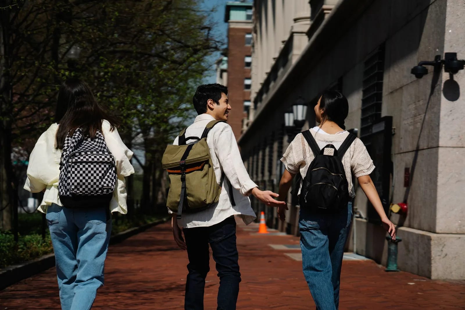 Three students carrying backpacks walking away along a brick path between campus buildings in spring