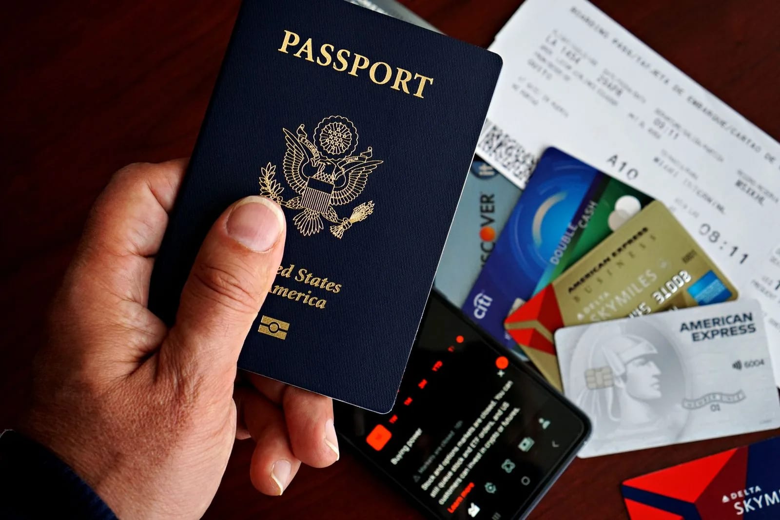 A hand holding a navy United States passport above a boarding pass, payment cards and a phone on a dark table