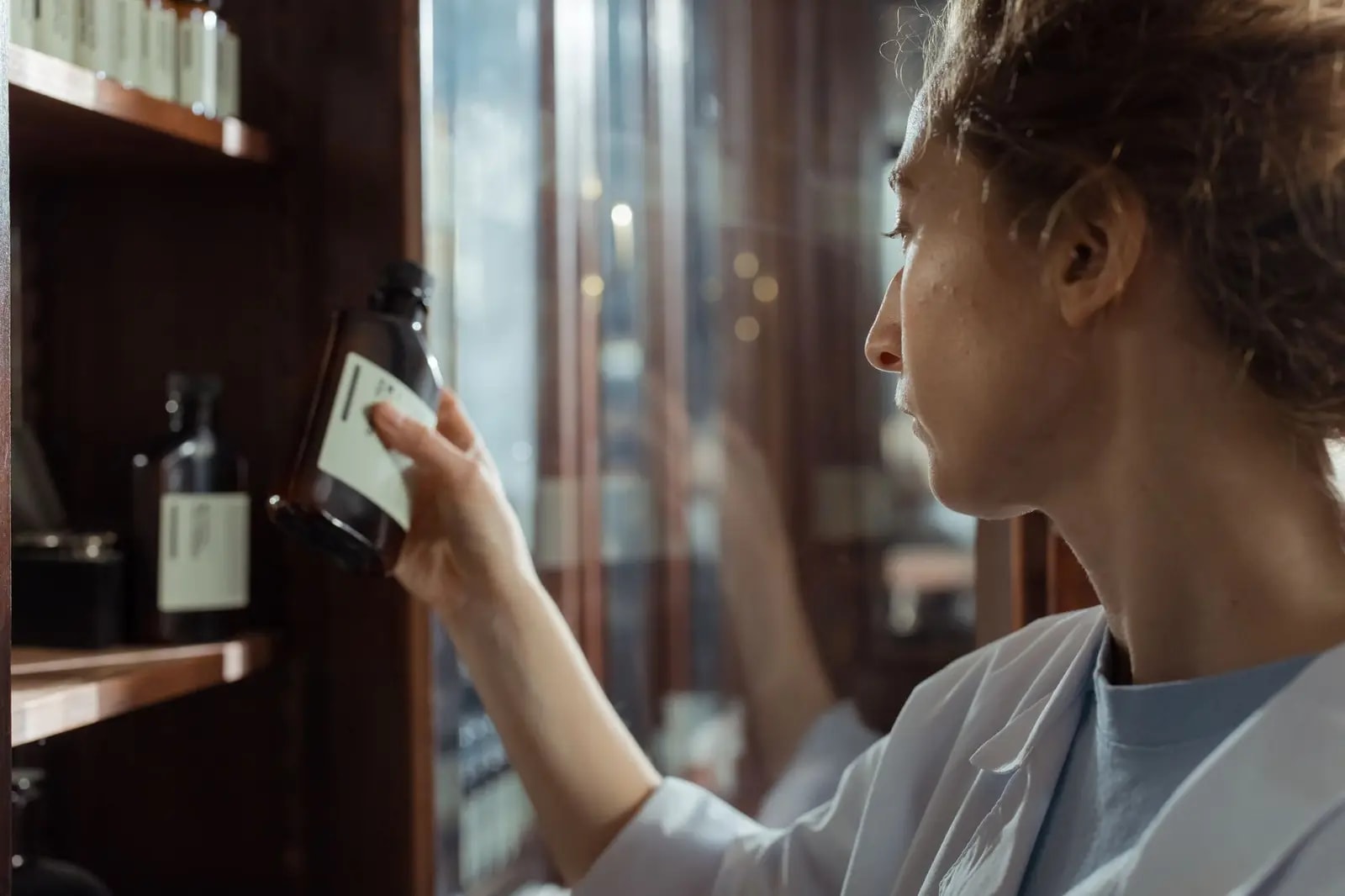 A pharmacist in a white coat lifting an amber bottle from a dark wooden dispensary cabinet