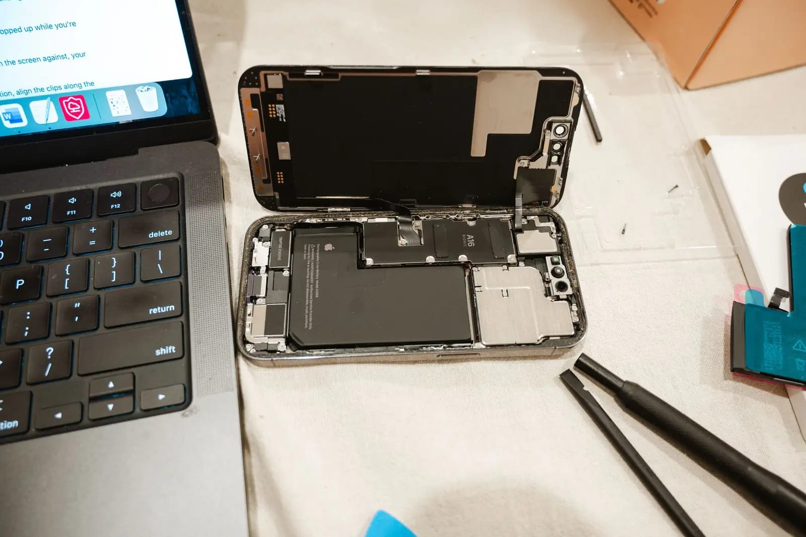 An opened smartphone with its back removed and battery exposed, beside repair tools and a laptop showing a guide