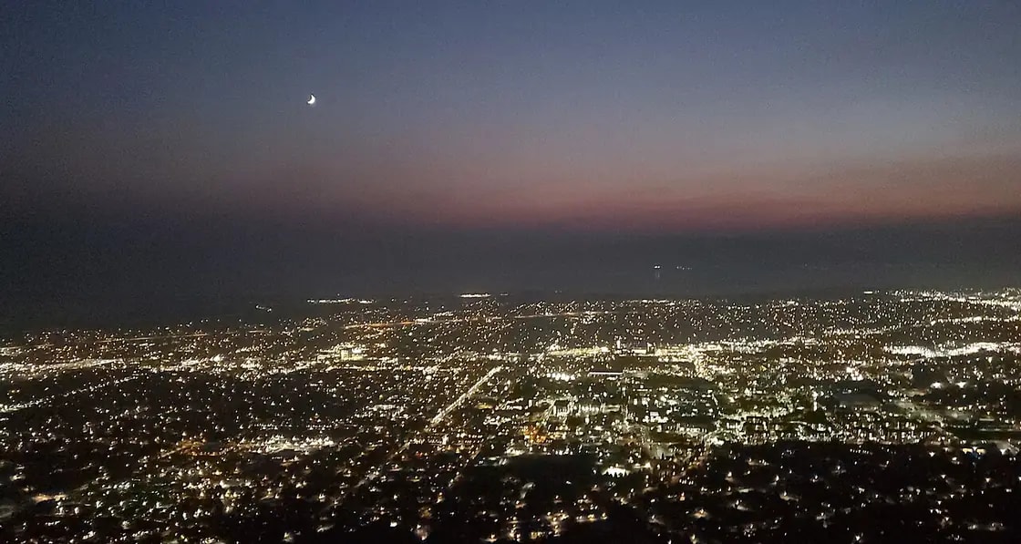 The lights of Provo at dusk seen from the top of Y Mountain, with a crescent moon above the afterglow on the horizon