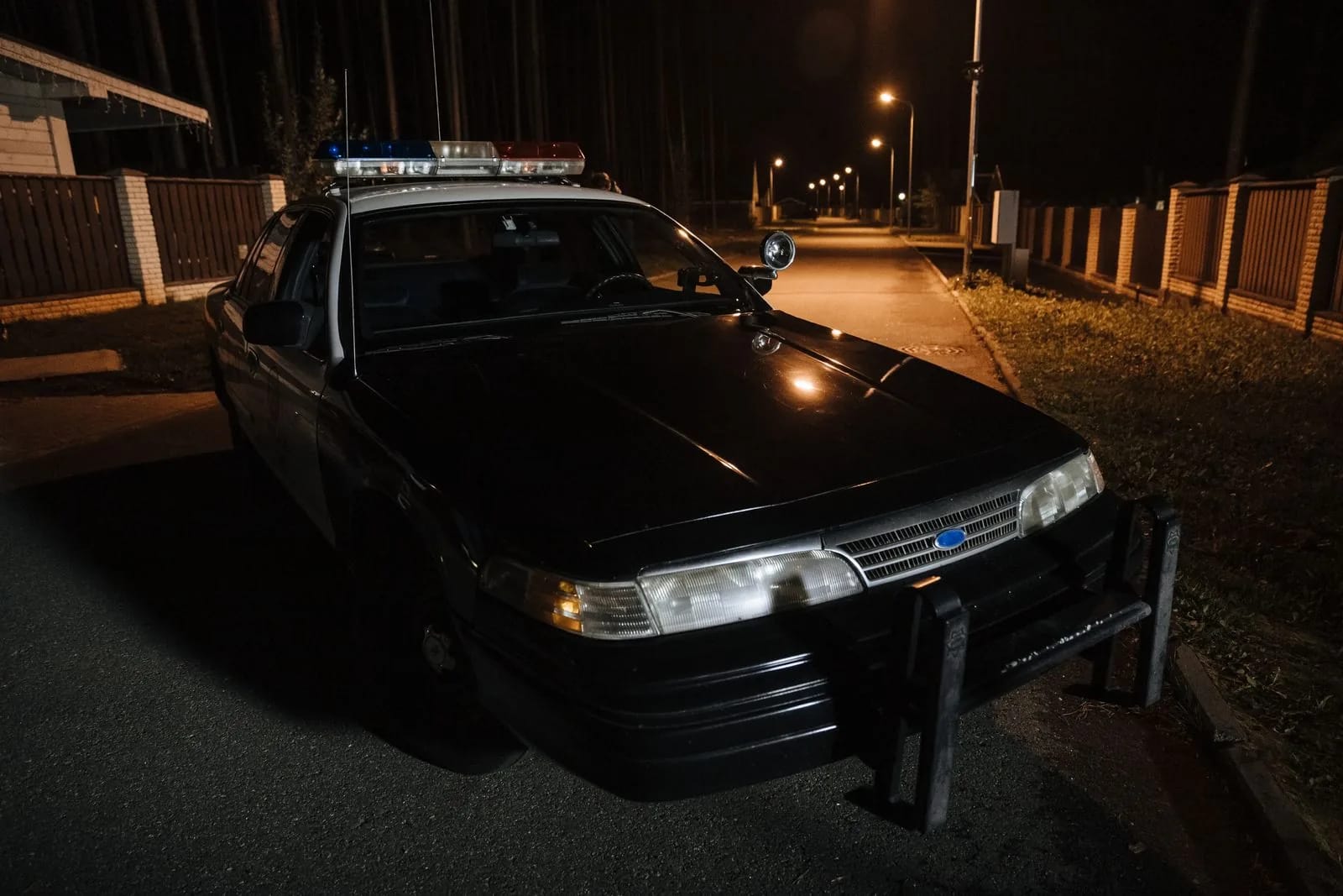 An older police sedan with a red and blue light bar and a front push bar, parked on an unlit street at night