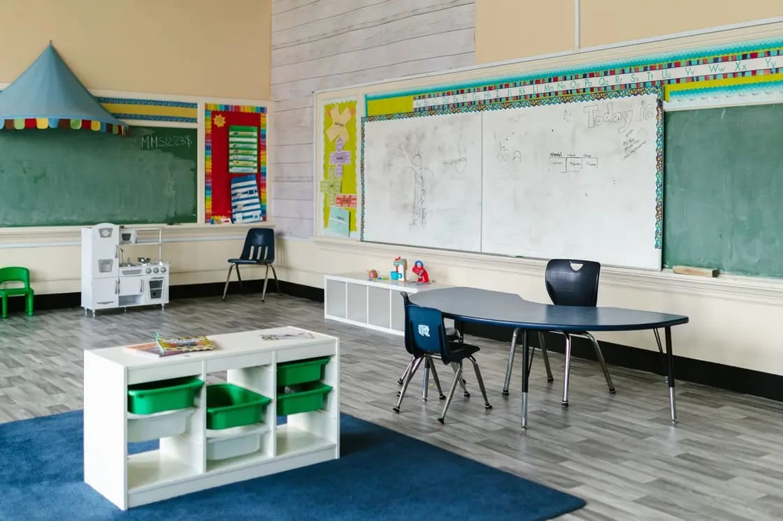 An empty preschool classroom with a kidney-shaped table, storage bins and an alphabet border above the whiteboard