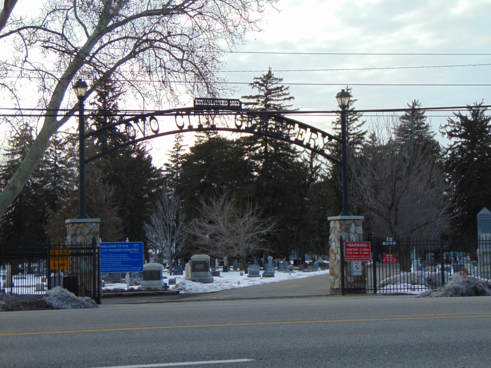 The stone and iron entrance arch of the Provo City Cemetery seen across South State Street on a winter afternoon, bare trees and snow patches beyond the gates