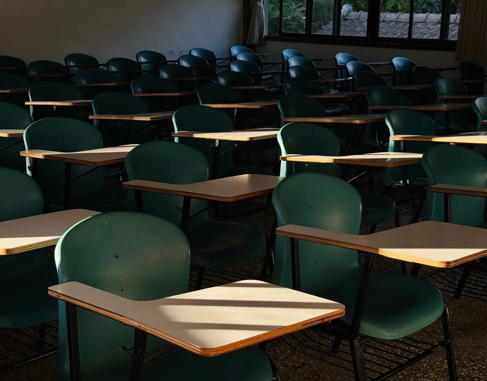 Rows of empty green tablet-arm school chairs with late sunlight falling across the desktops