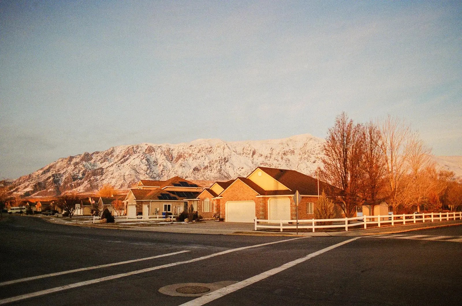 A quiet residential street of low houses with snow-covered mountains rising behind them