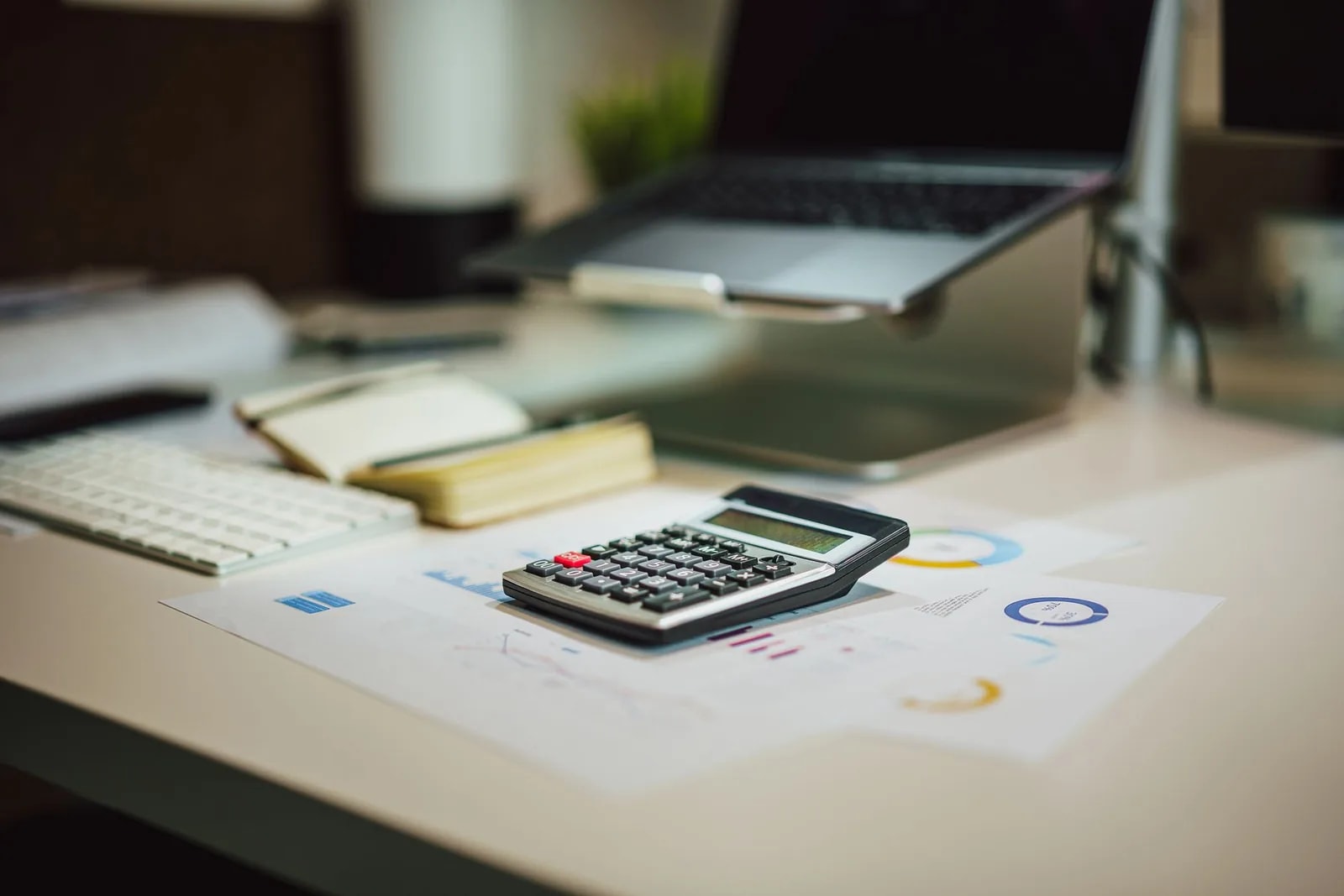 A calculator, notepad and printed financial charts on a desk beside a laptop