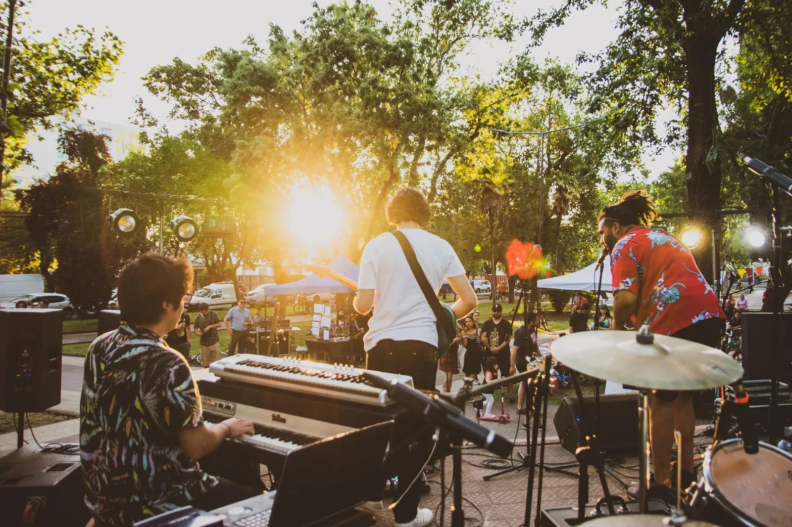A band playing keyboards, guitar and drums on an outdoor stage under trees in late afternoon sun