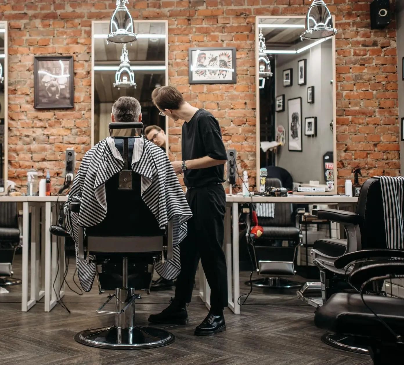 A barber working on a caped client in a brick-walled shop lined with mirrors and empty chairs