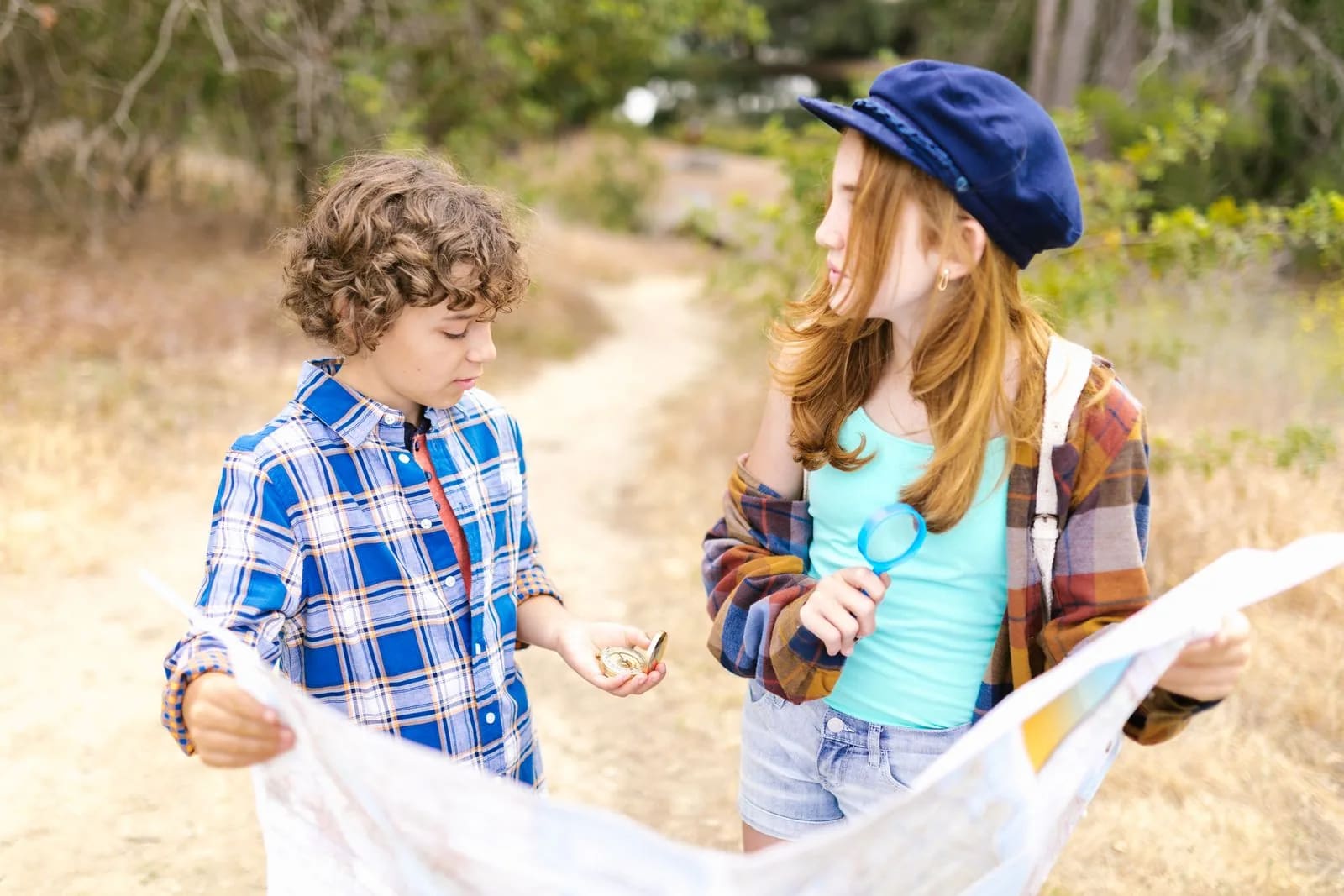 Two children on a dirt path holding an open paper map, one with a compass and one with a magnifying glass