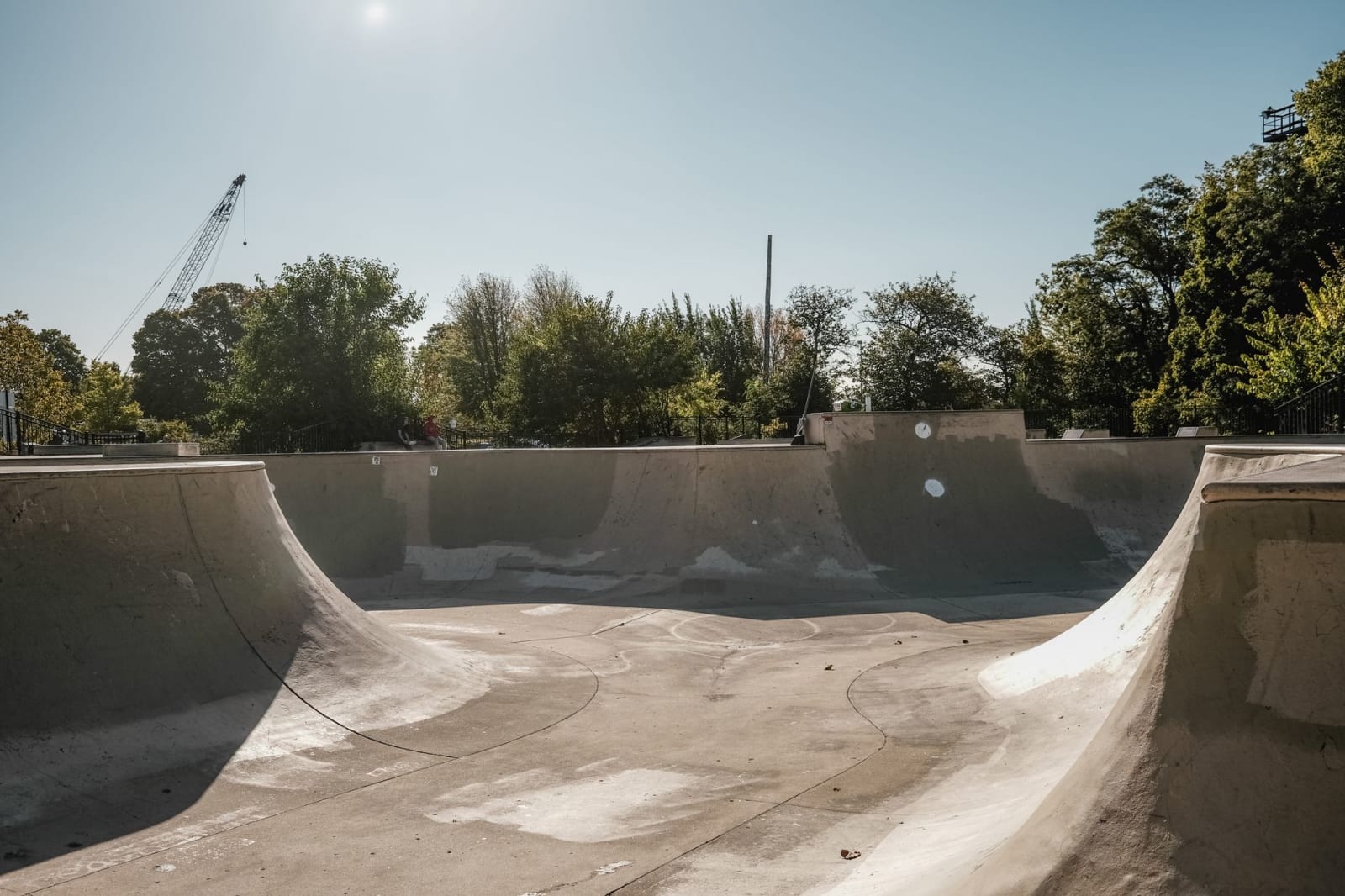 An empty concrete skate bowl under a clear blue sky, with trees along the far edge
