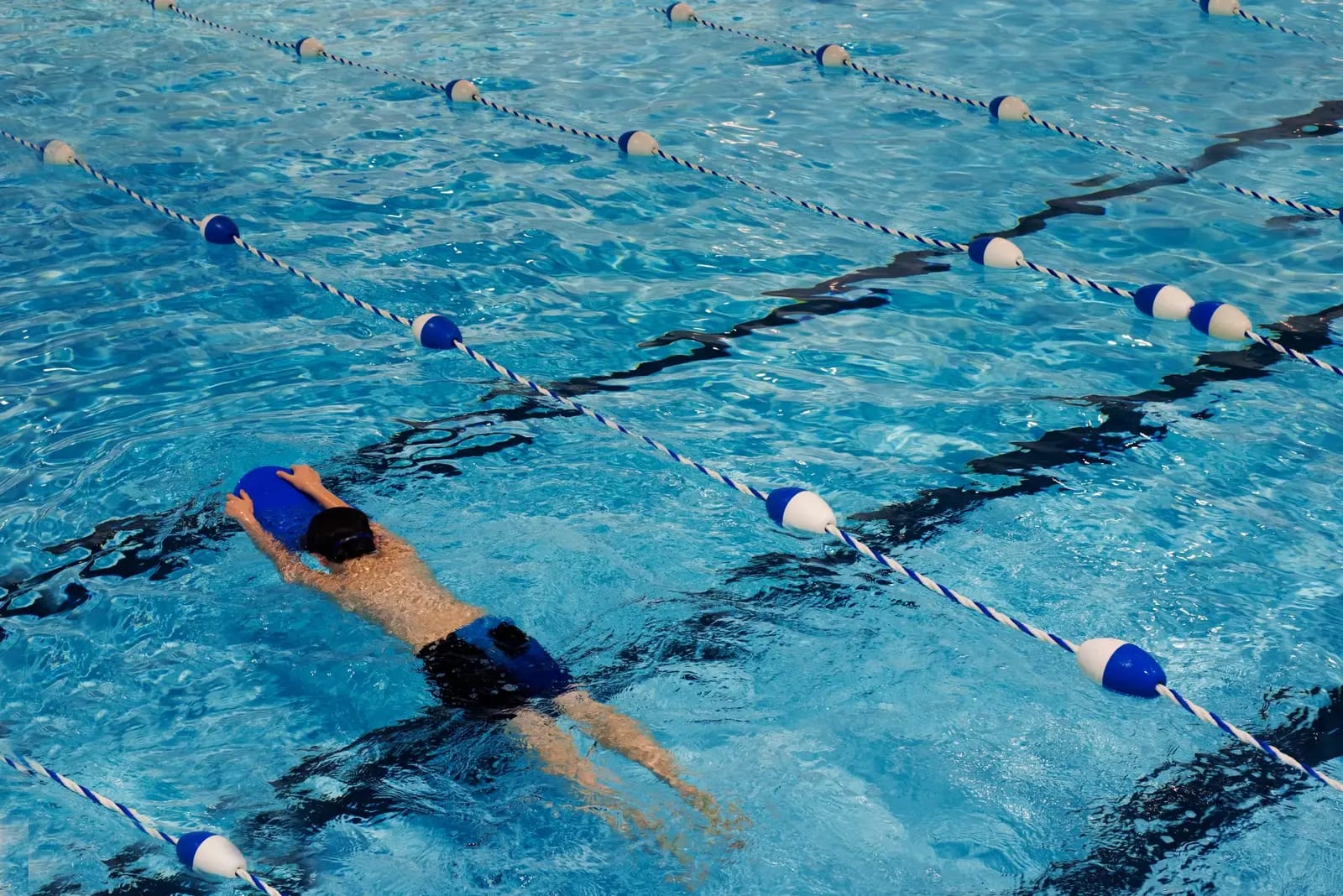 Seen from above, a swimmer kicking behind a blue kickboard between the lane ropes of an outdoor pool