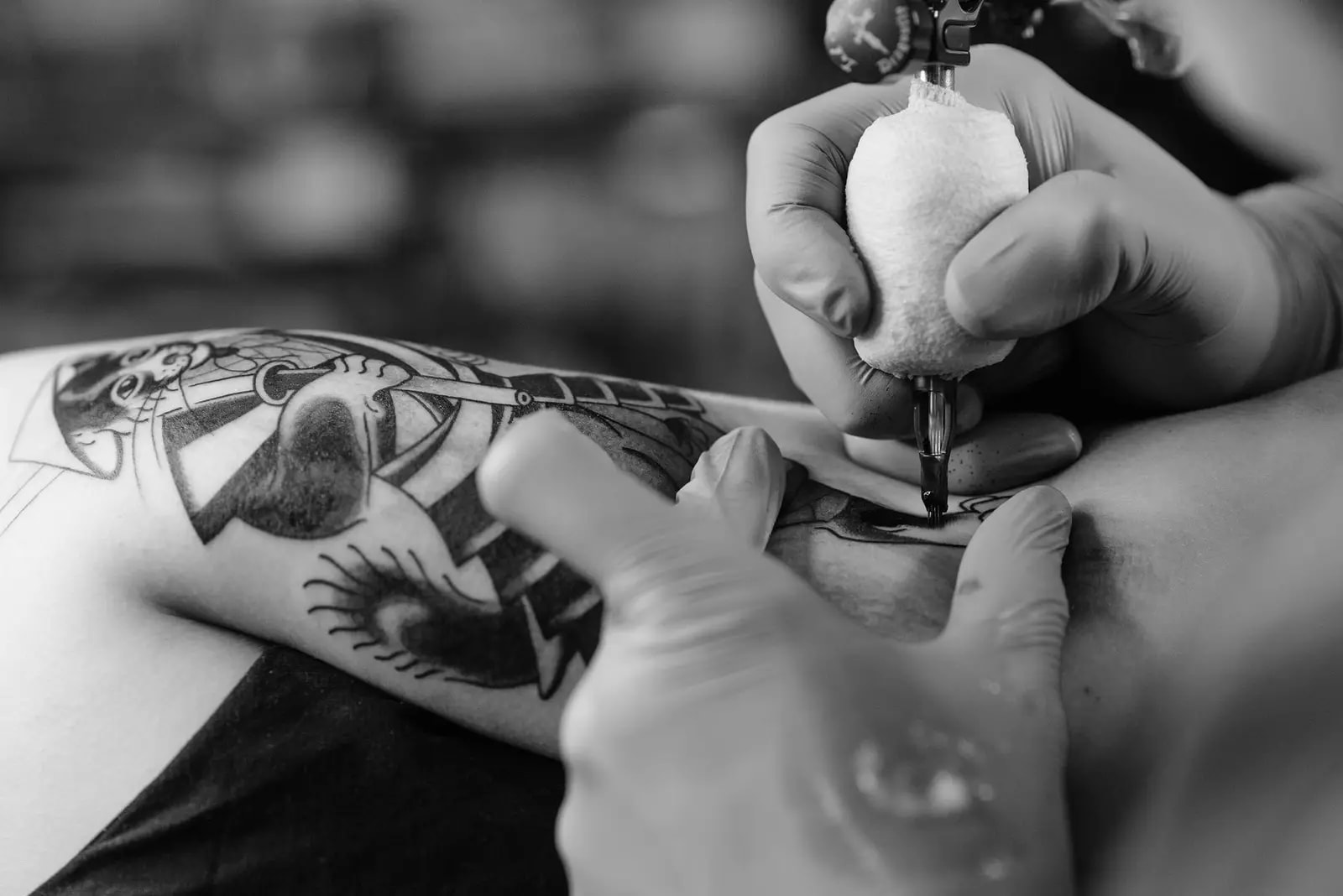Gloved hands working a tattoo machine on a forearm already covered in bold traditional designs