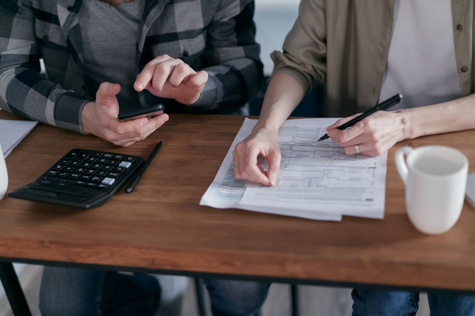 Two people at a wooden table filling out a printed tax form, with a calculator and a mug beside them