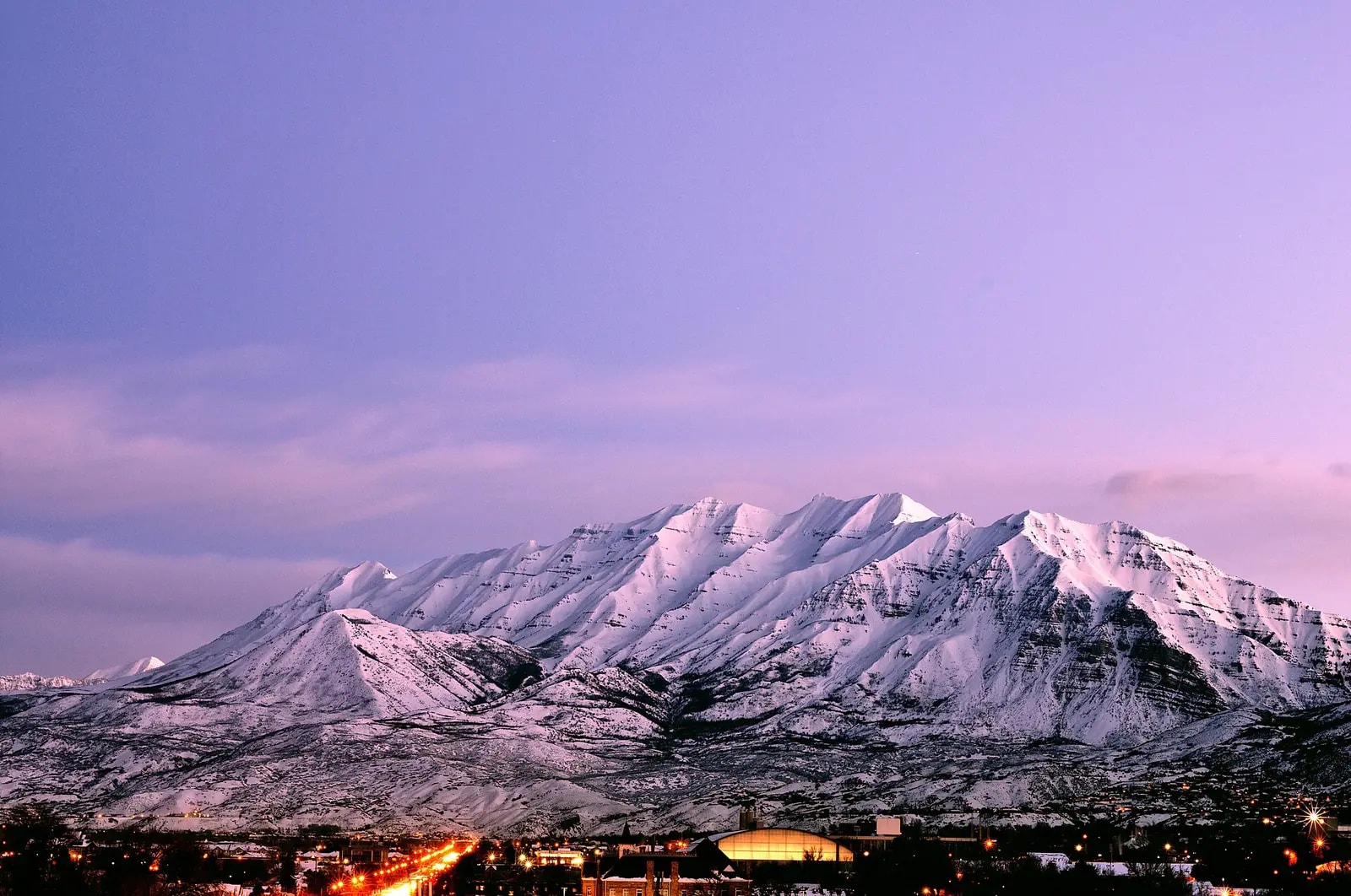 The long snow-covered ridgeline of Mount Timpanogos at dusk under a violet sky, with the lights of the valley strung along its base