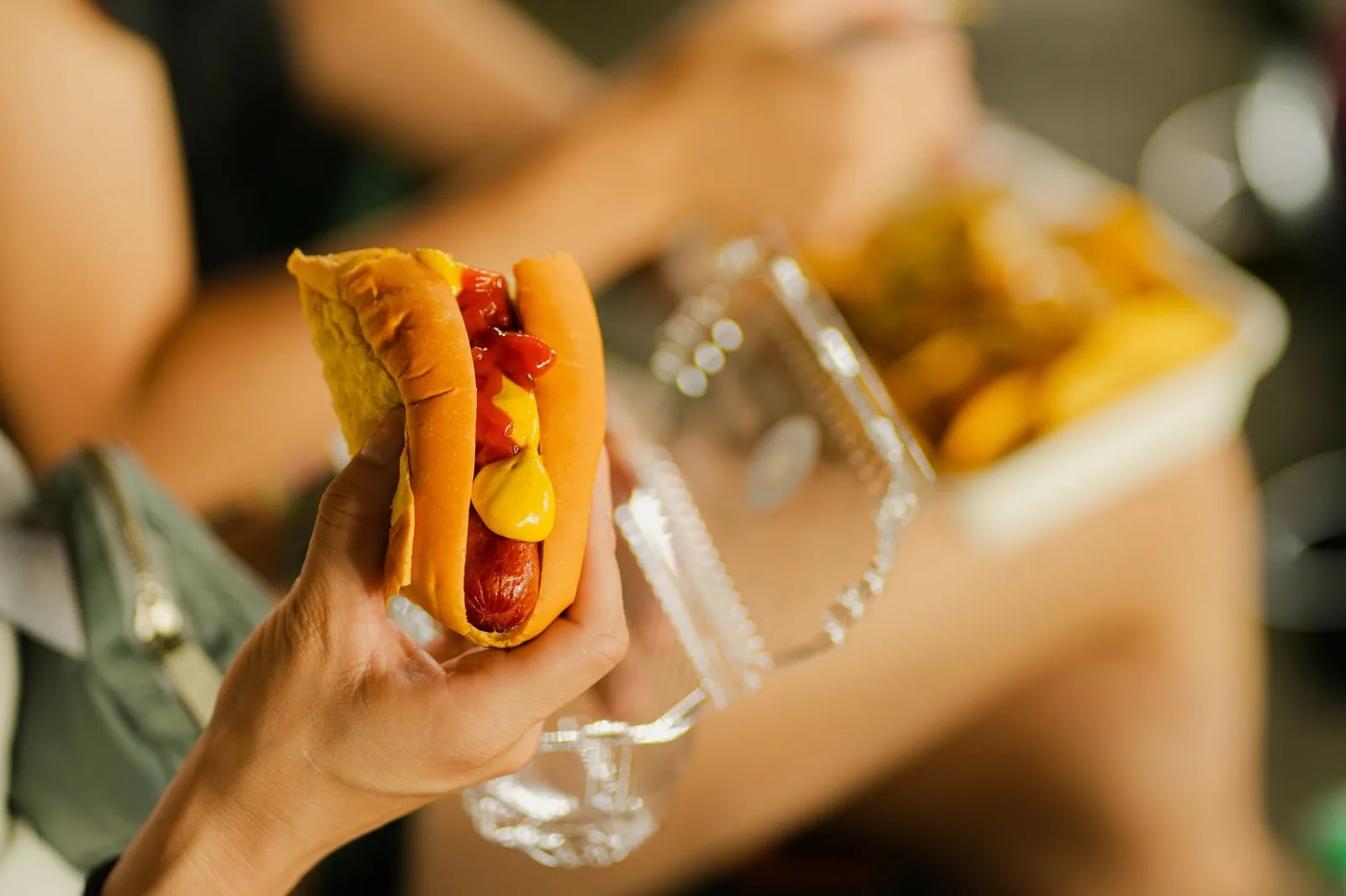A hand holding a hot dog with mustard and ketchup, a tray of fries behind