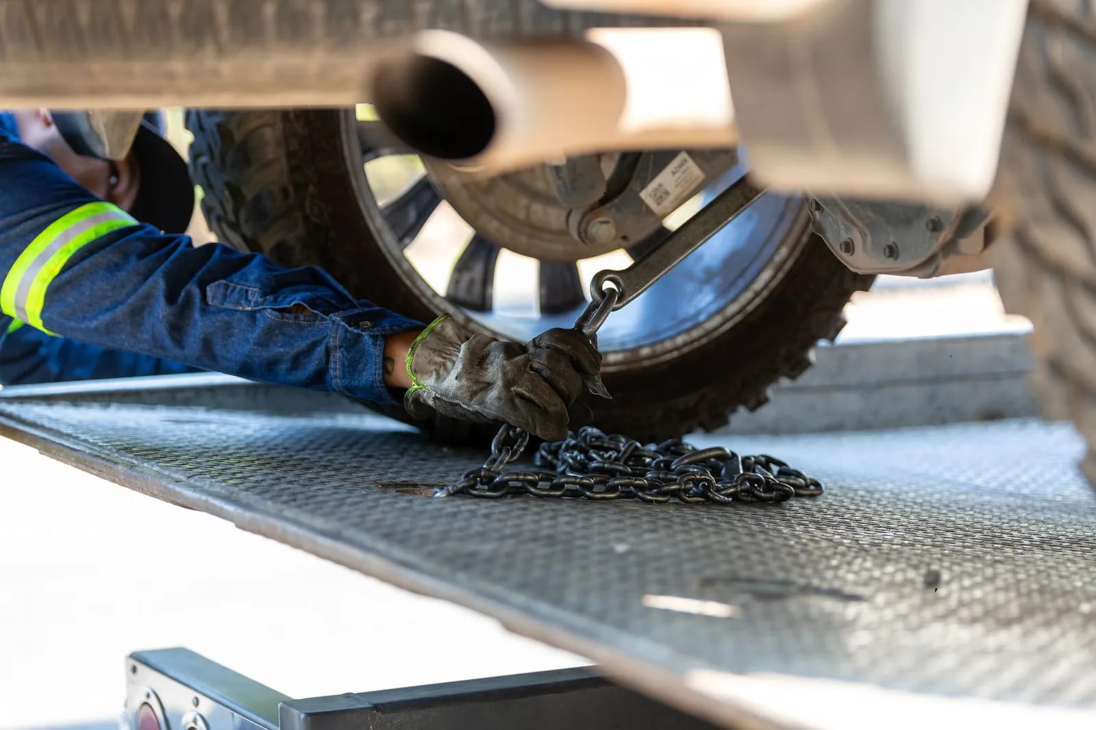 A worker in high-visibility clothing securing a chain around a vehicle wheel on a flatbed tow truck deck