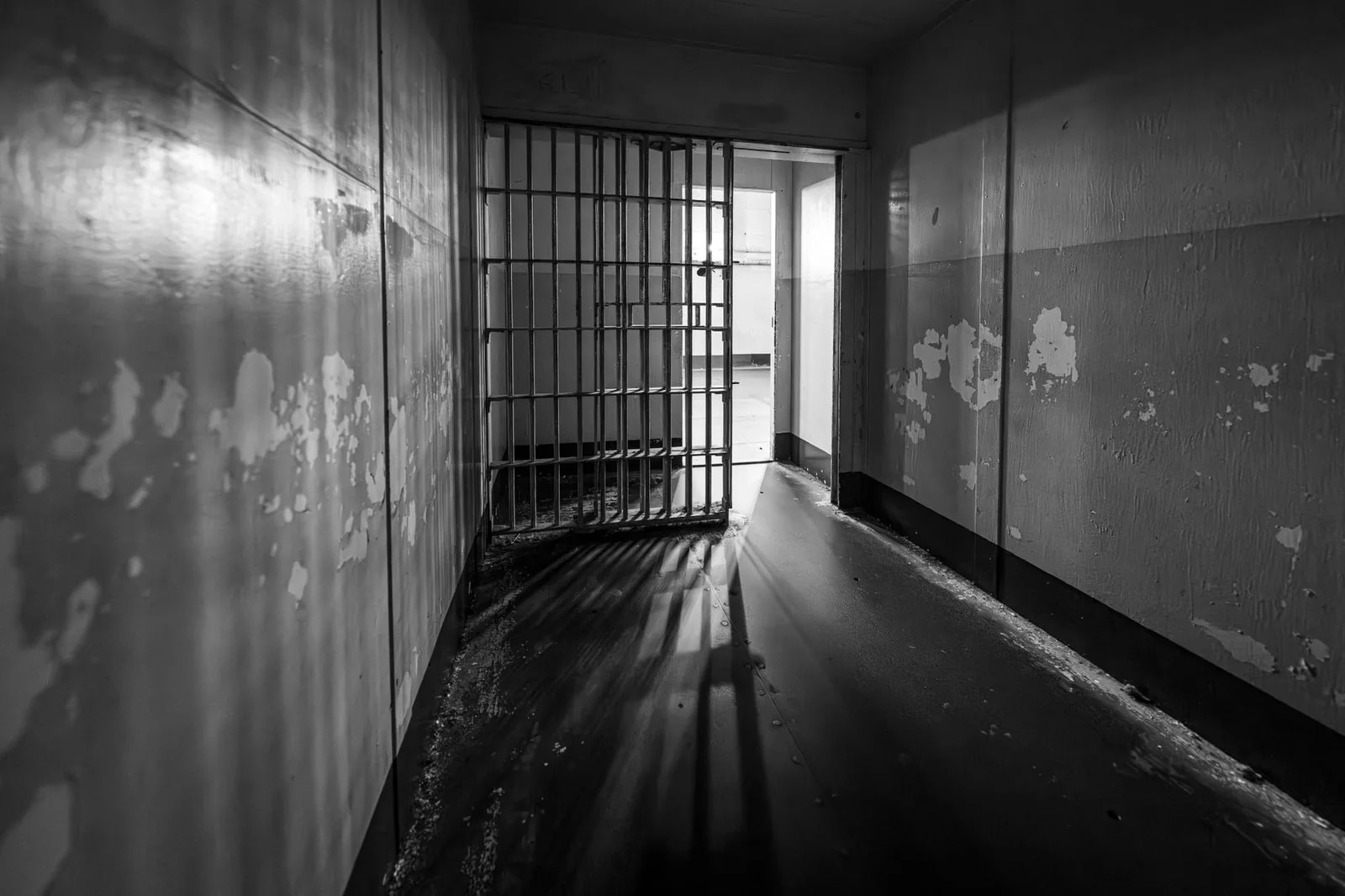 A barred cell gate at the end of a dim corridor with peeling walls, in black and white