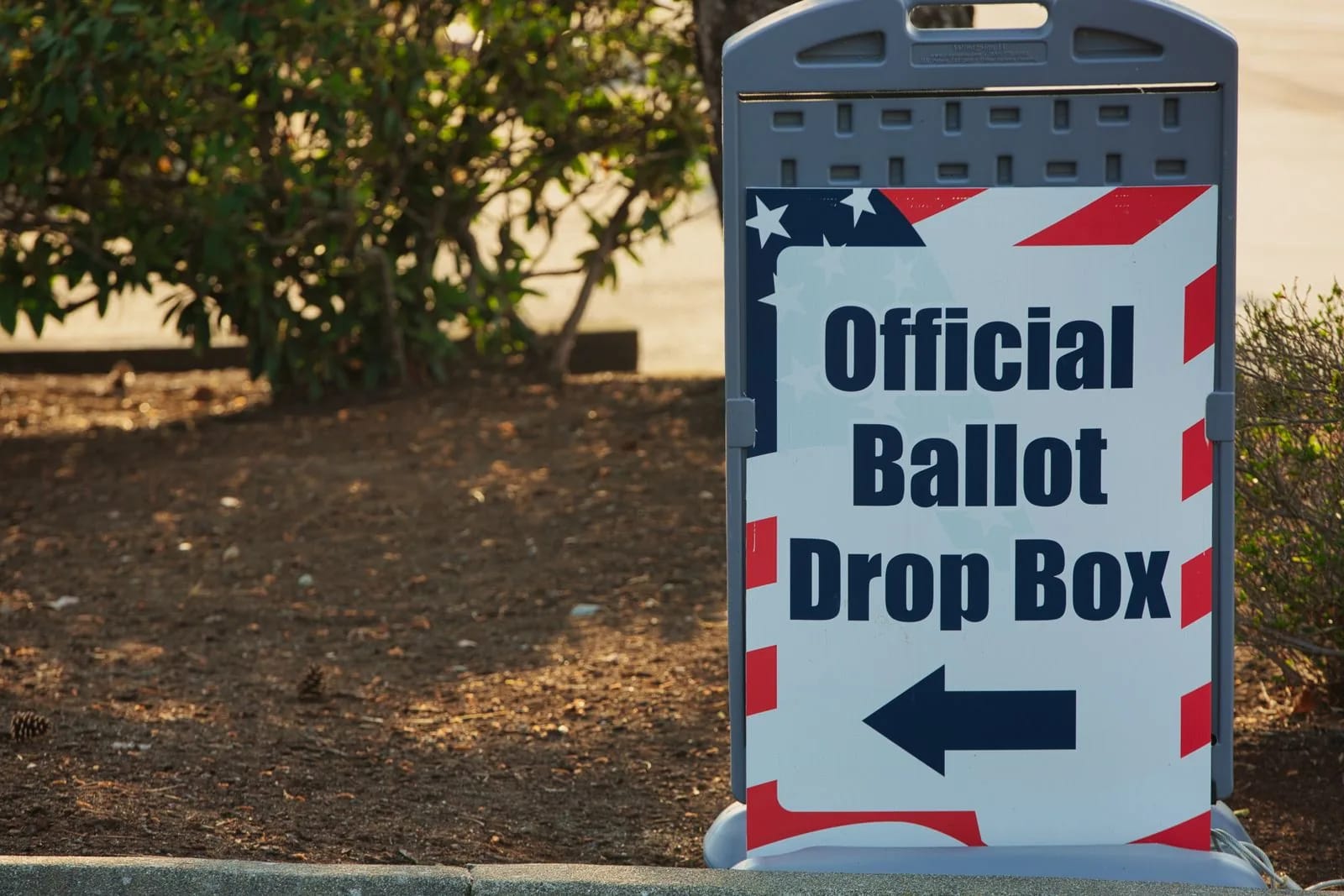 A stars-and-stripes sign reading Official Ballot Drop Box with an arrow, standing on a curb beside a shrub