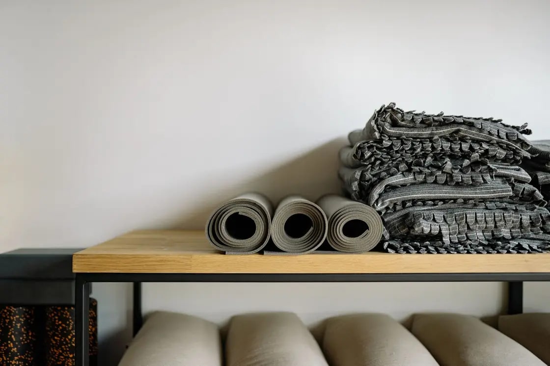 Rolled mats and folded blankets on a wooden studio shelf above a row of bolsters