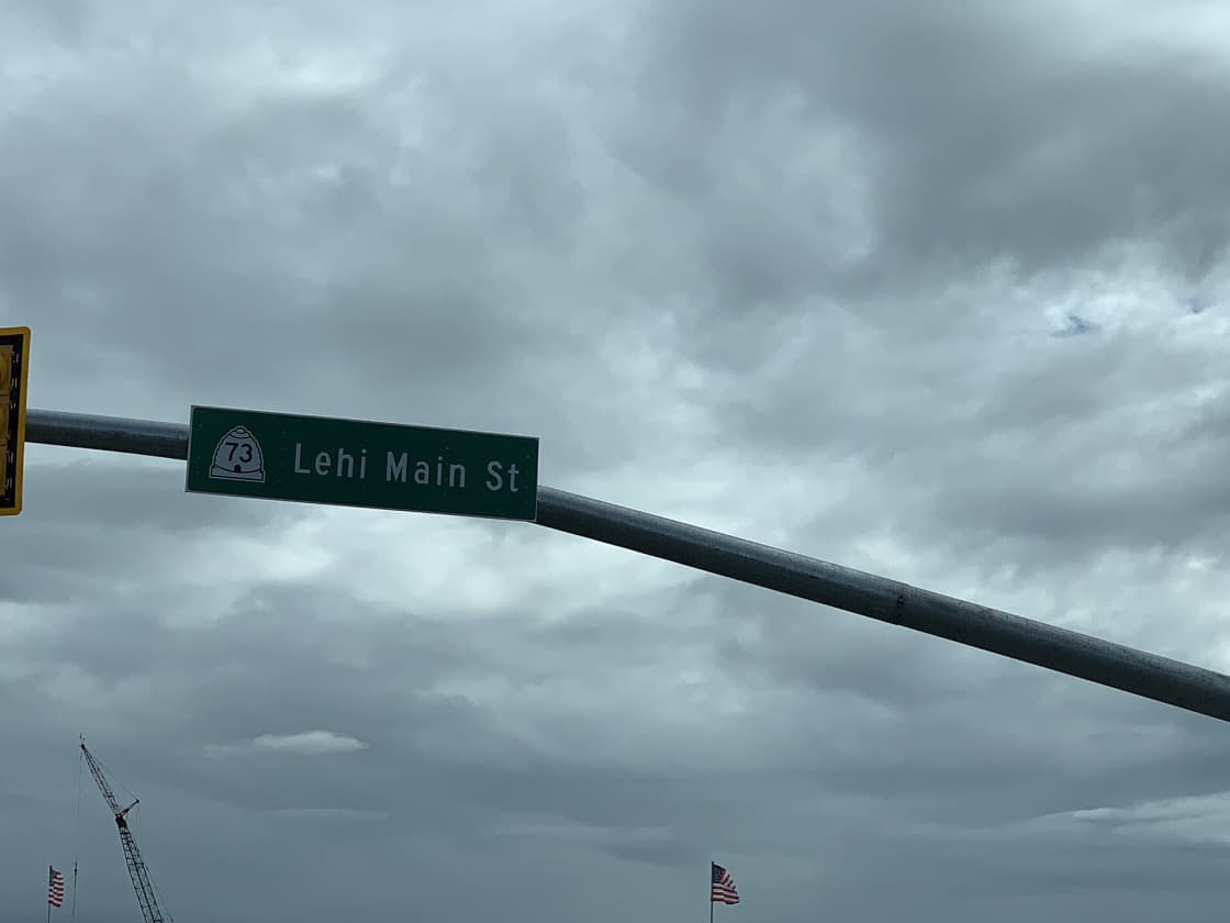 A green street sign reading Lehi Main St on its mast arm against a heavy gray sky, with a construction crane and a flag behind