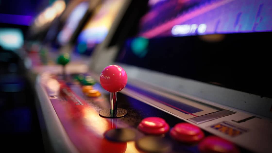 A pink arcade joystick and red buttons on a lit control panel in a darkened arcade