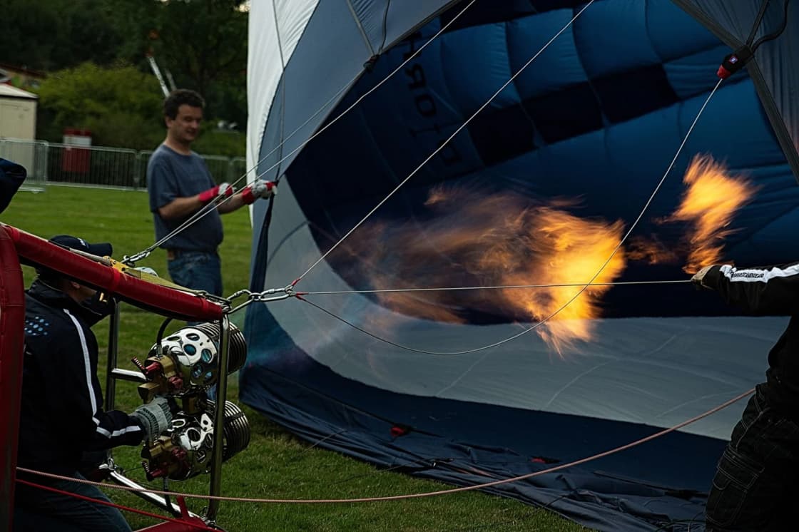 A ground crew inflating a hot air balloon with a burner flame while holding the lines on a grass field
