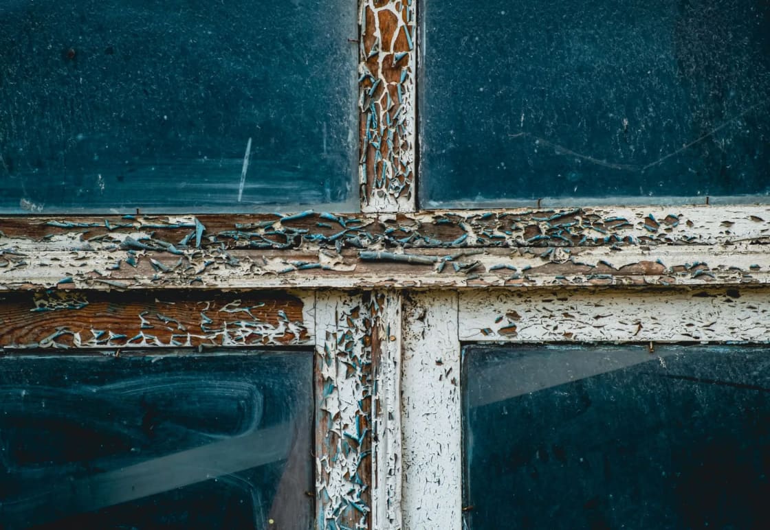 Thickly cracked and peeling white paint on the wooden bars of an old window, with bare wood showing through