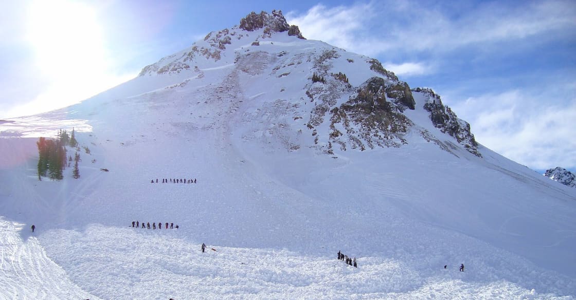 Rescuers spread in probe lines across a wide avalanche debris field below a steep mountain peak