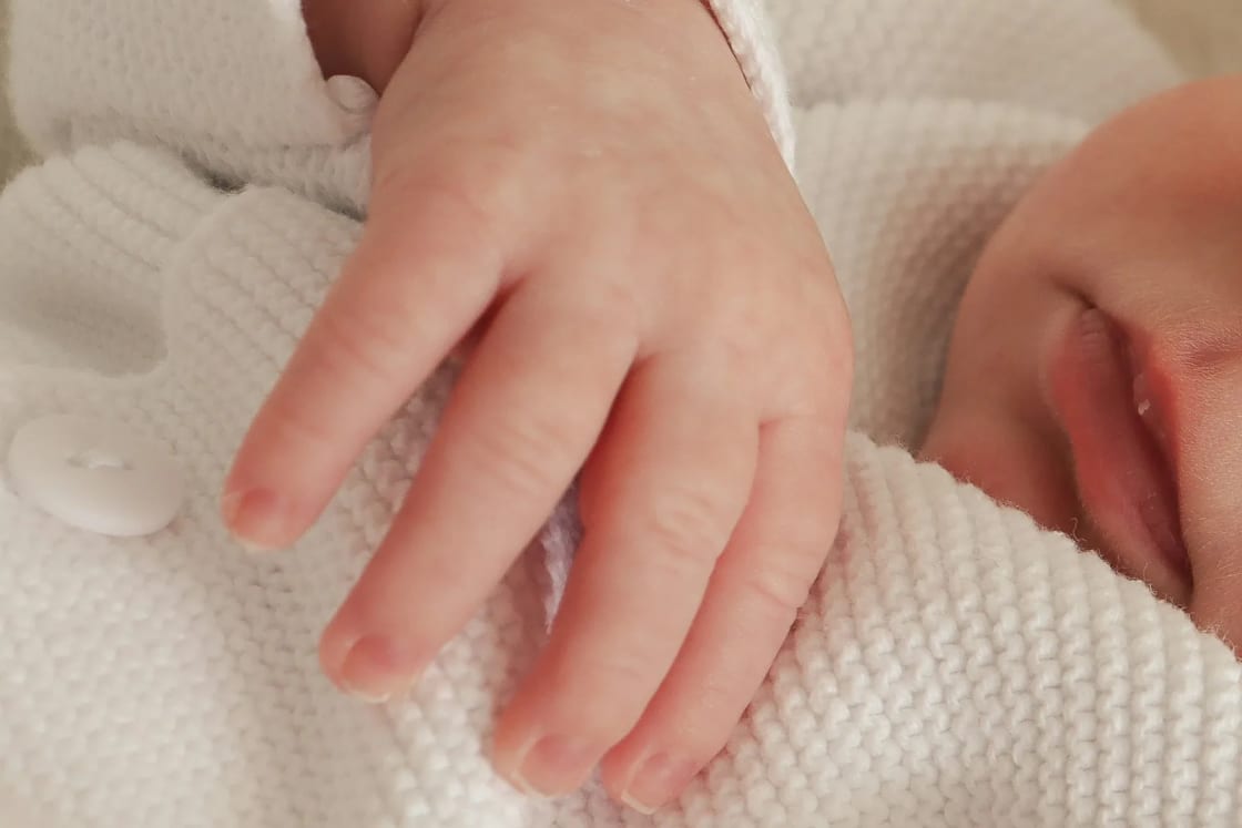 A sleeping newborn's small hand resting on a soft white knit outfit, the kind of white clothing many families choose for a baby blessing