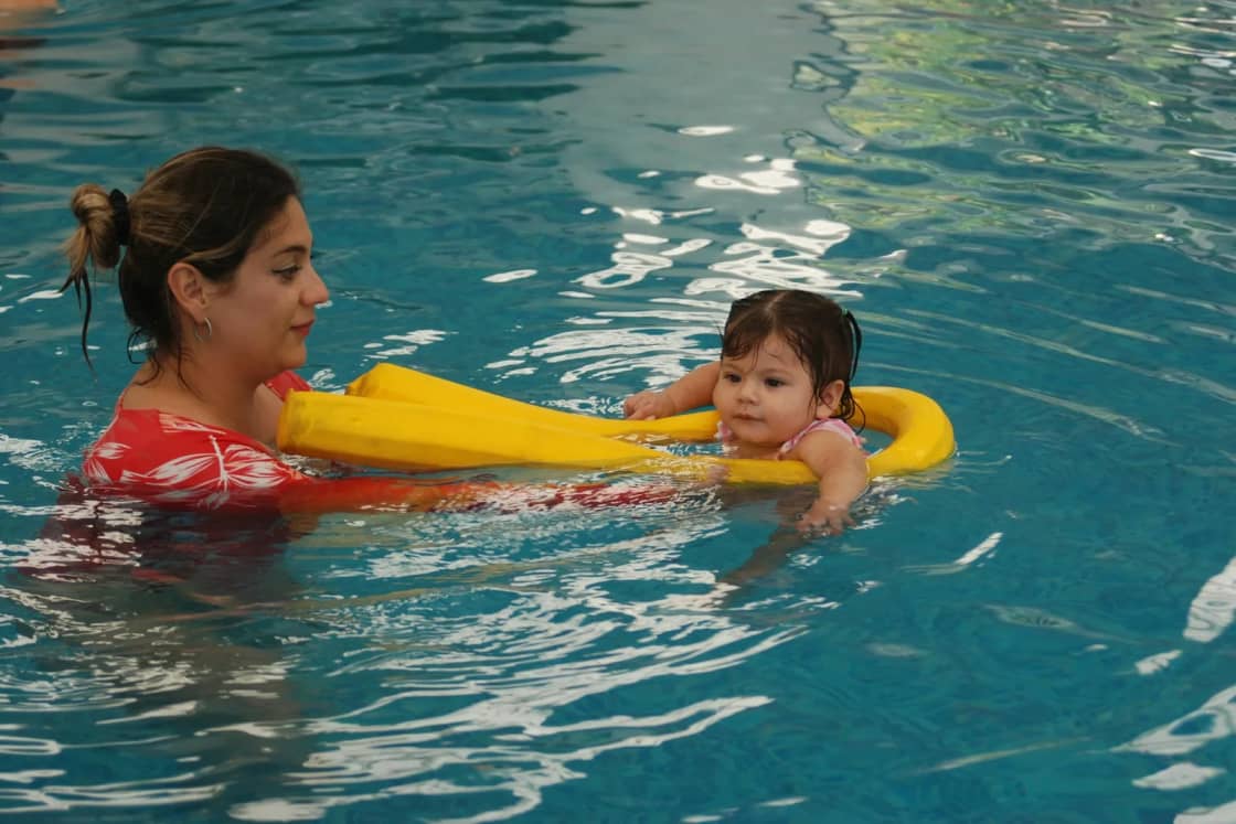 A mother in an indoor pool guides her baby, who floats on a yellow foam noodle, during a parent-and-child swim session.
