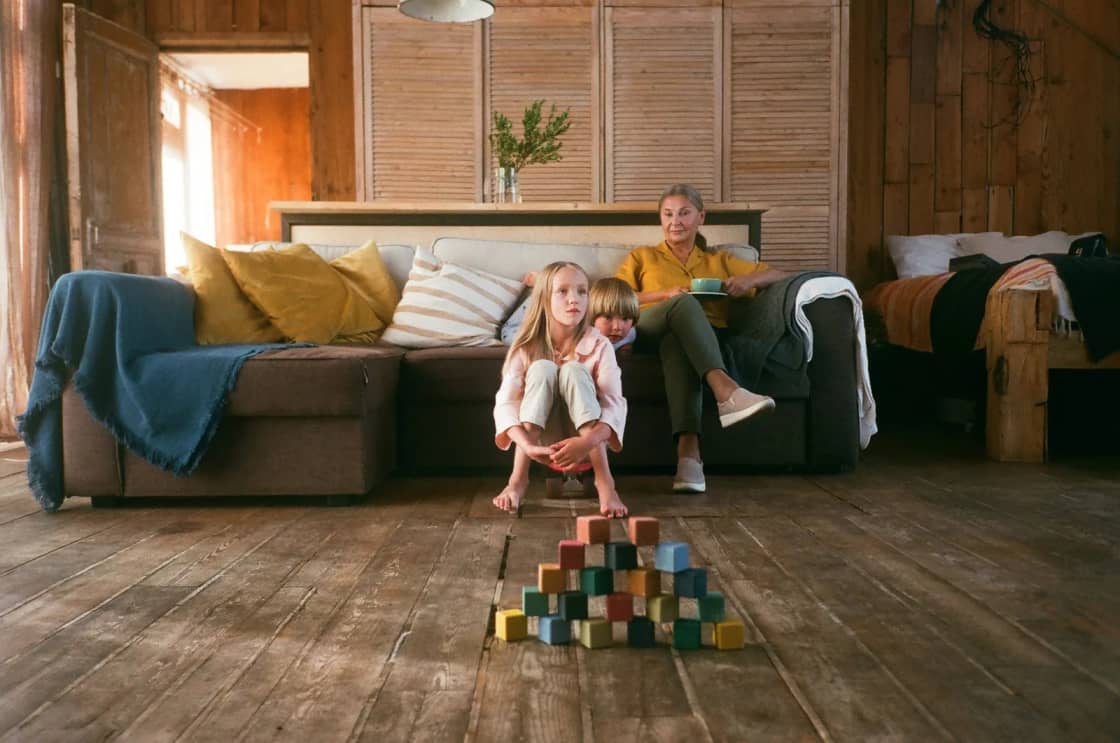 A young child stacking colored wooden blocks into a pyramid on a wooden floor while an adult sits watching from a low couch