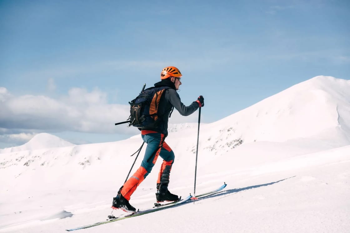 A skier in an orange helmet and backpack skins uphill on touring skis across a bright, wind-sculpted snow slope under a blue sky