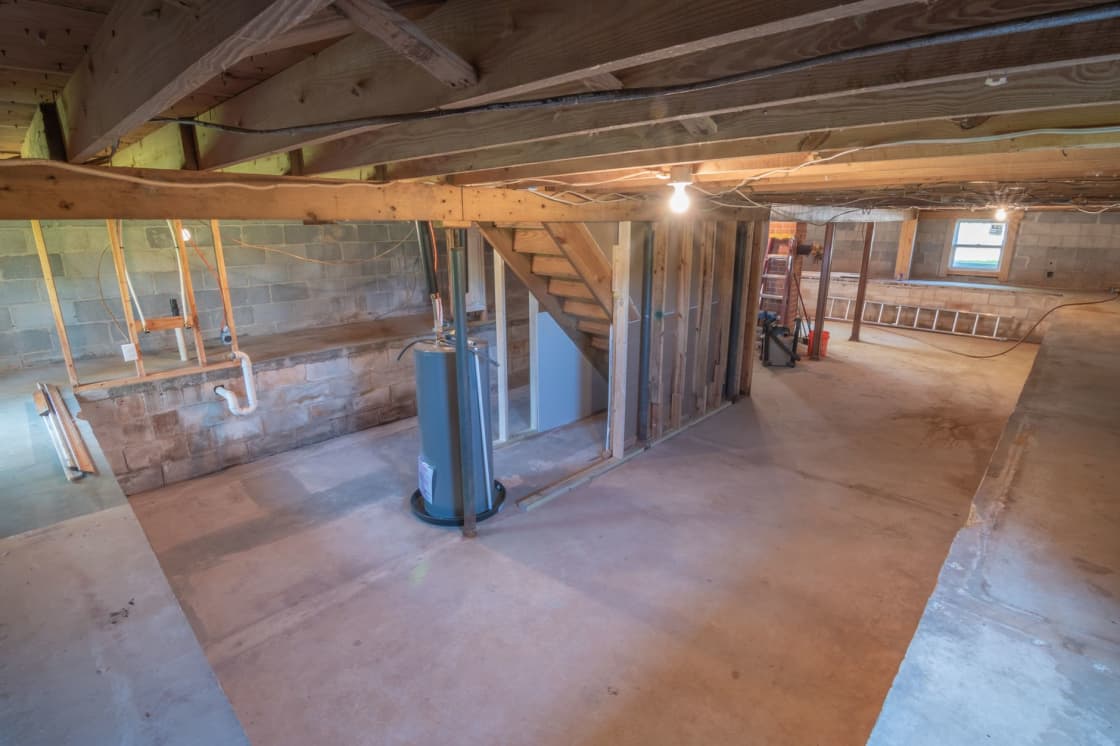 An unfinished basement with a bare concrete floor, concrete block walls, exposed floor joists overhead, a partition of bare wood studs and a water heater standing on a drip pan