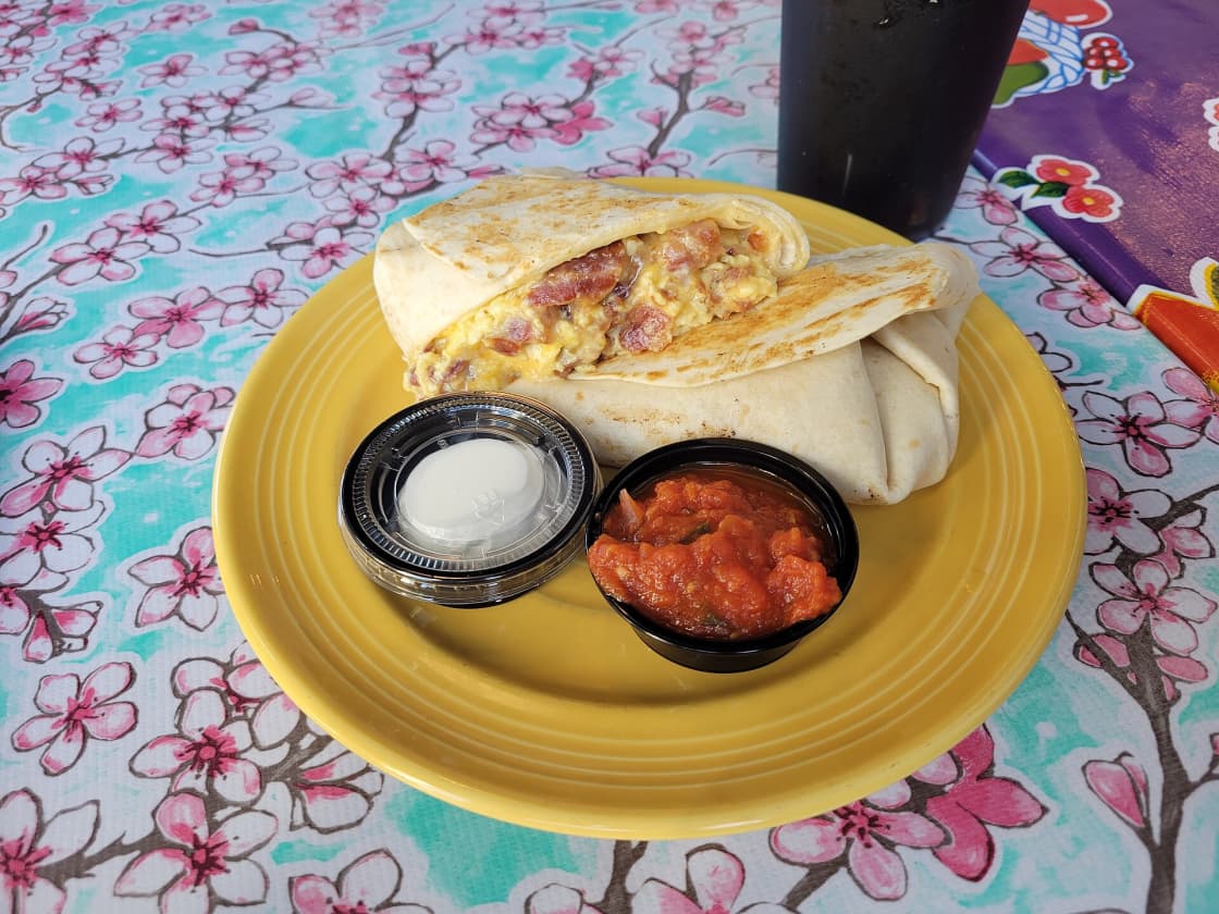 A breakfast burrito cut in half on a yellow plate showing egg, bacon and cheese, with salsa and sour cream alongside