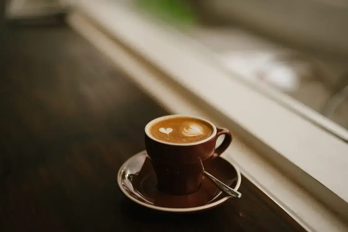 A cappuccino with latte art on a saucer beside a sunlit window