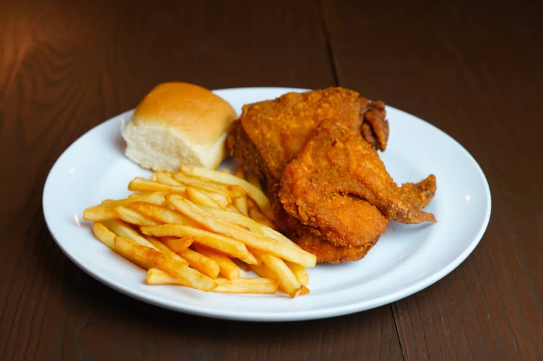 A white plate holding bone-in fried chicken pieces, a pile of french fries and a soft dinner roll on a dark wooden table