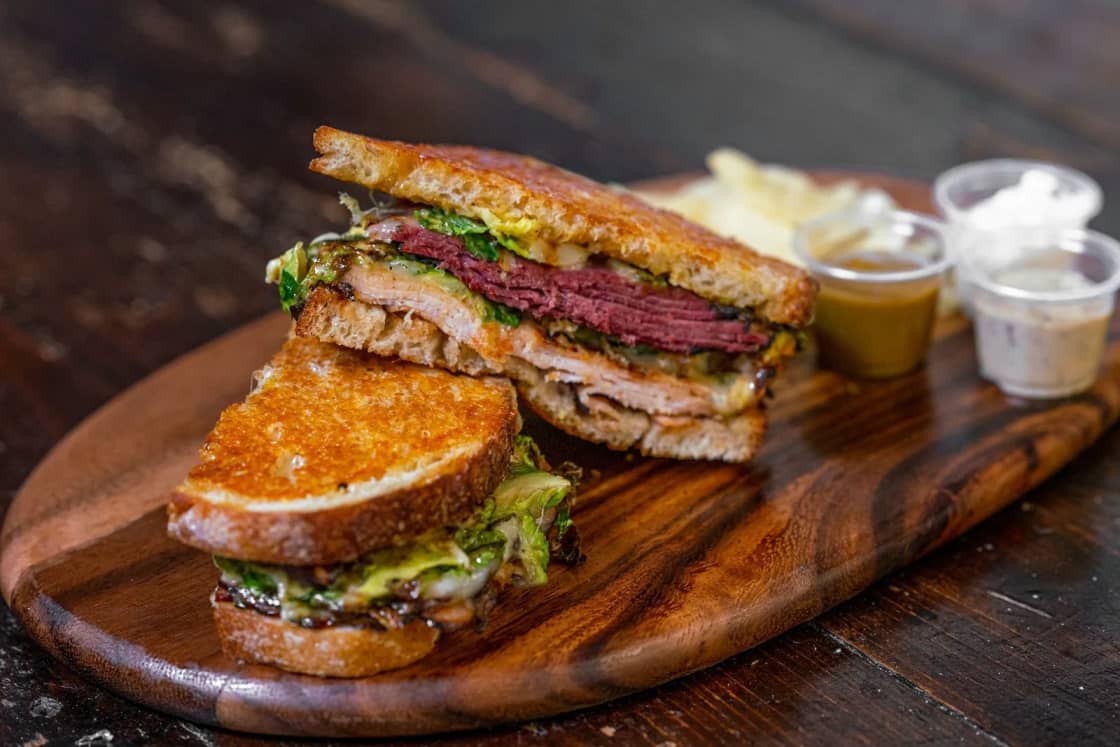 A toasted deli sandwich cut in half and stacked on a dark wooden board, layered with pastrami, turkey, melted cheese and greens, with chips and two dipping sauces alongside