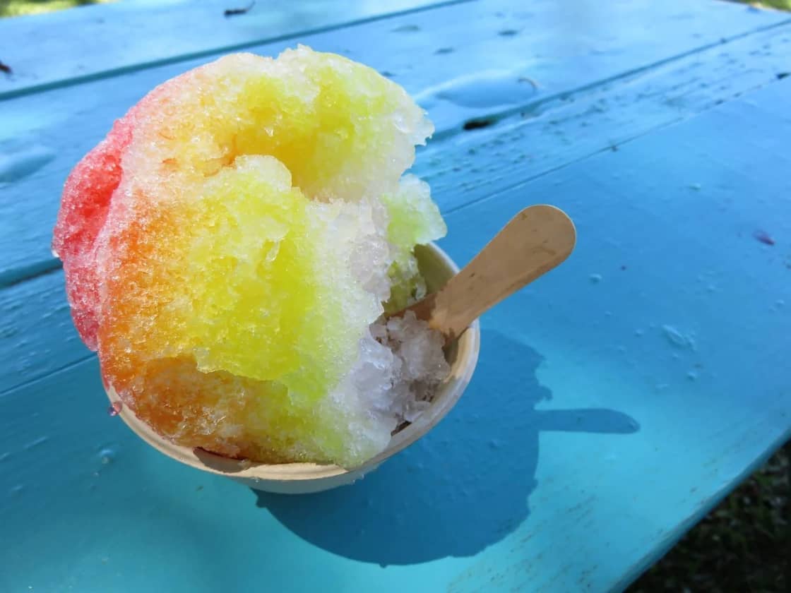 A paper cup of rainbow shave ice with a wooden spoon on a weathered turquoise picnic table