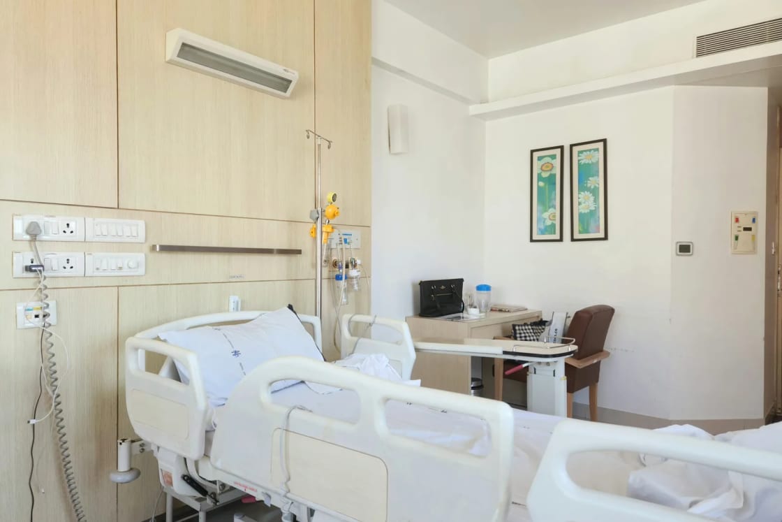 An empty, sunlit hospital room with an adjustable bed, side rails, wall-mounted medical outlets and a small desk and chair.