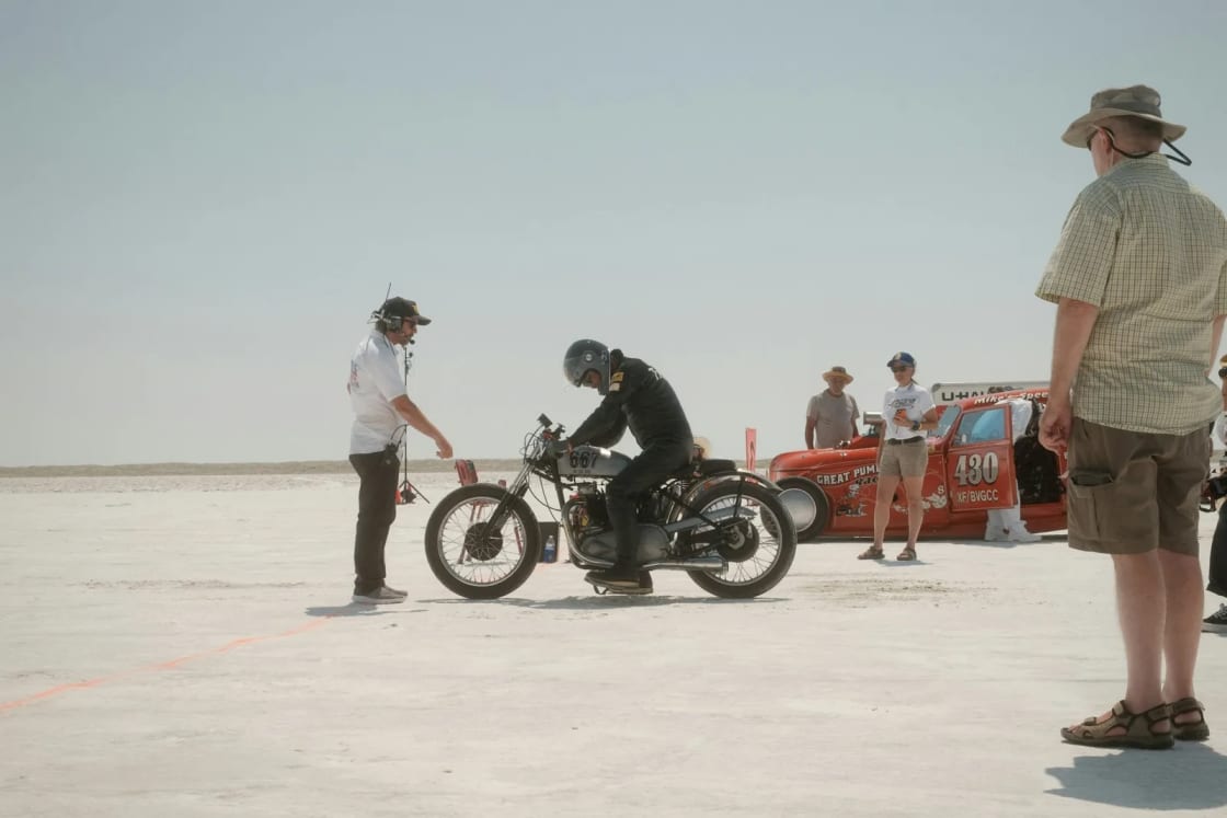 A rider on a vintage motorcycle waits on the white salt of the Bonneville Speedway as crew and spectators stand near an orange race car.