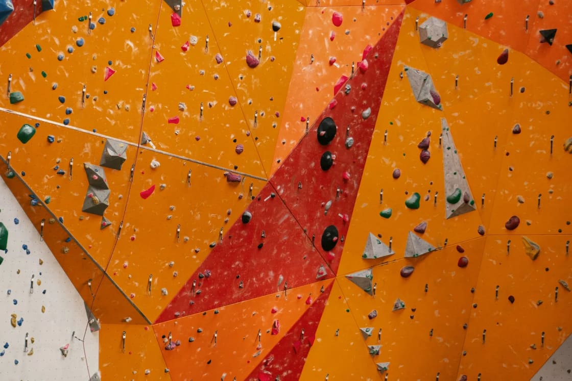 A towering indoor climbing wall painted in orange, red and yellow facets and covered in colorful holds, with a climber small at its base