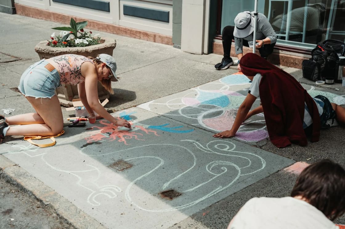 Several artists kneeling on a sunny downtown sidewalk in front of shop windows, drawing large, colorful chalk pictures in squares on the concrete