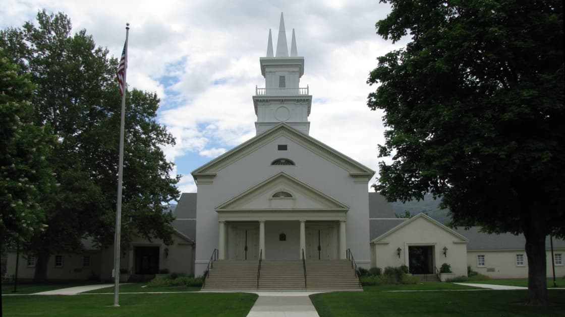 The white Bountiful Tabernacle on Main Street, with its tall steeple and spires, columned portico and broad front steps, a flagpole and shade trees on the lawn under a cloudy sky